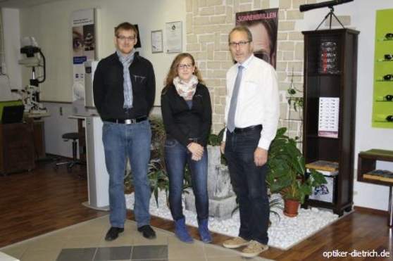 Drei Personen stehen im modernen Optikerladen vor einer Wand mit Brillenregal und Pflanzen.